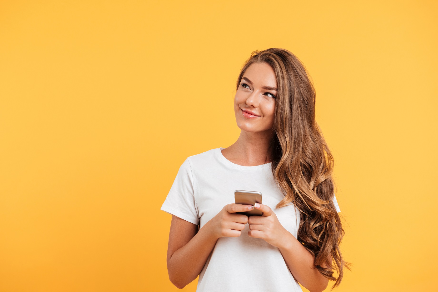 Photo of cheerful cute beautiful young woman chatting by mobile phone isolated over yellow wall background. Looking aside.