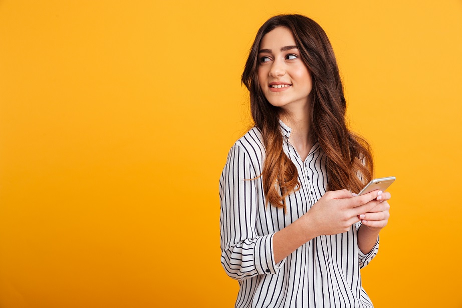 Portrait of a pretty young girl holding mobile phone while standing and looking away isolated over yellow background